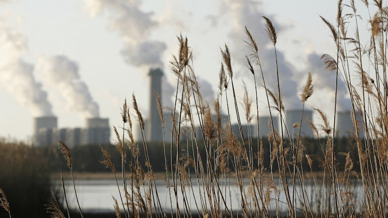 Grass is seen in the foreground with a lake beyond and long plumed from cooling towers on the other side of the lake.