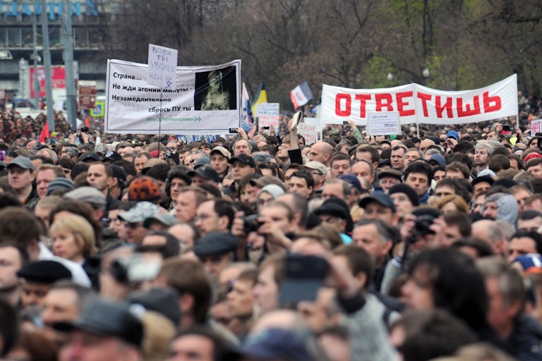 A crowd of protesters outdoors facing slightly left, with some signs in Russian visible in the back.