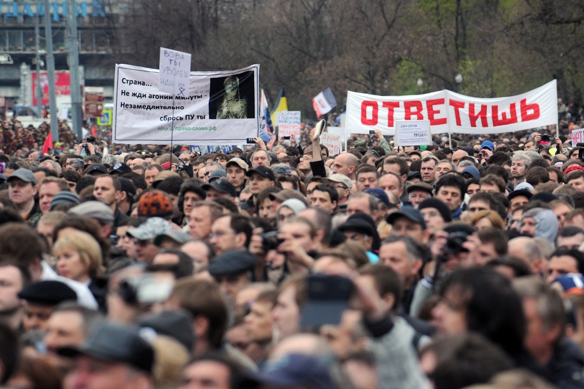 A crowd of protesters outdoors facing slightly left, with some signs in Russian visible in the back.