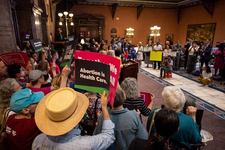 Abortion rights advocates protest indoors. One person in the foreground holds a sign reading "Abortion is health care." Another in the background reads "Bans off our bodies."
