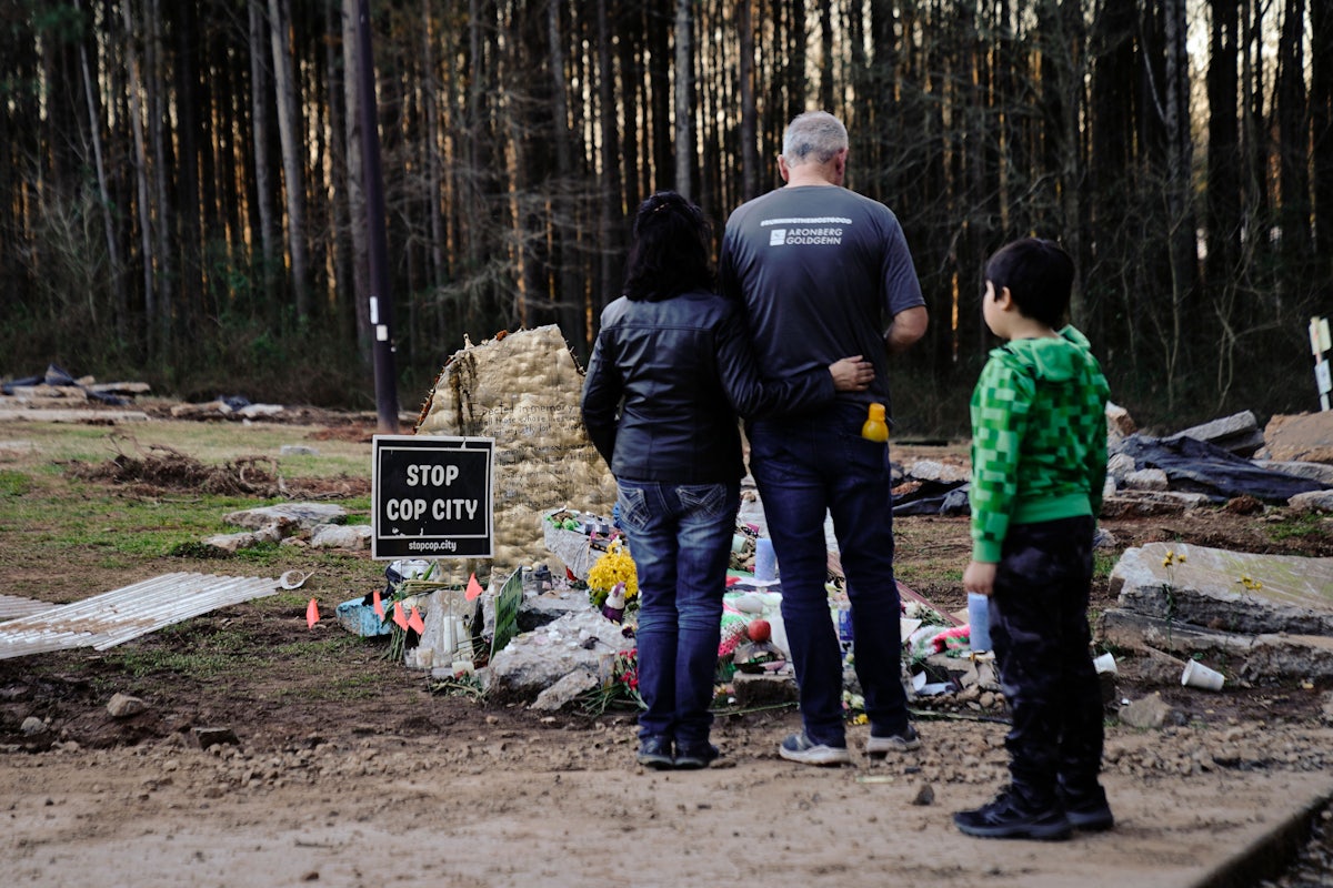 Three people stand at the site of a memorial, next to a sign reading "Stop Cop City."