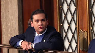 Representative George Santos leans over a railing in the House chamber and looks directly at the camera