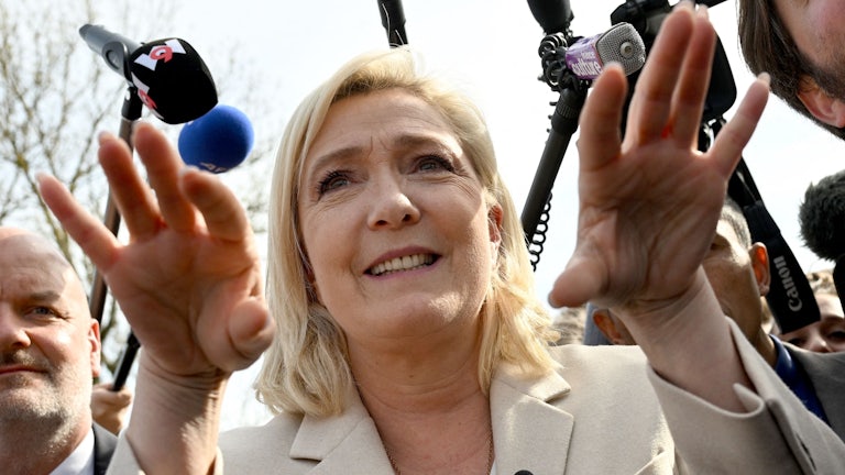 Marine Le Pen gesticulates while speaking, with microphones in her face.
