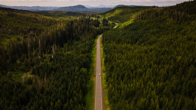 Aerial view of a truck driving through a heavily forested road with mountains in the background. 