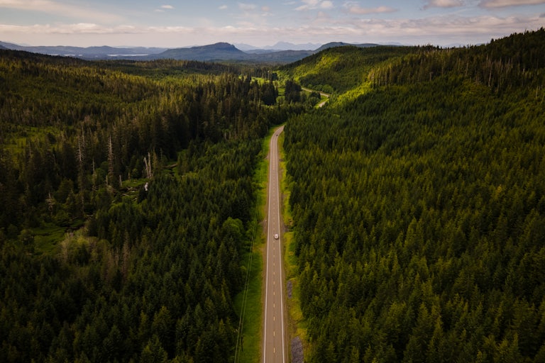Aerial view of a truck driving through a heavily forested road with mountains in the background. 