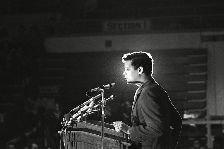 A black-and-white photograph of Julian Bond standing before a lectern equipped with microphones.