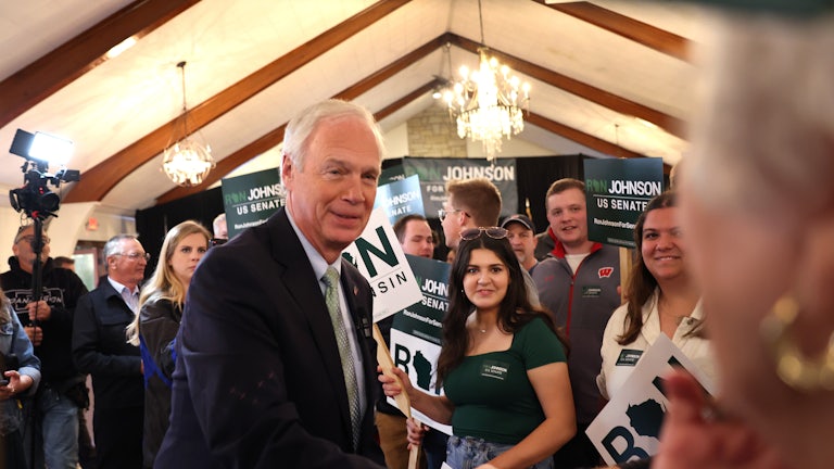 Ron Johnson shakes someone's hand as supporters stand around him with signs that read "Ron Johnson for US Senate." A camera is in the background.