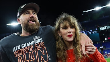 Travis Kelce throws his arm around Taylor Swift's shoulders. They're standing on the football field smiling. Kelce wears a cap and a shirt that says "AFC Champion Chiefs Are All In." Taylor Swift wears a red sweater and red lipstick.
