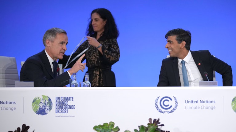 Two men sit behind a table, talking, as a woman walks behind them.