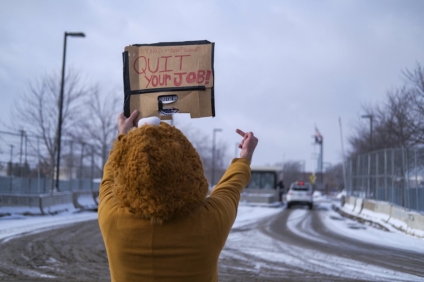 A protester stands next to the road holding a sign that reads HEY NAZI, COLD, SCARED? QUIT YOUR JOB while giving an unmarked vehicle the middle finger
