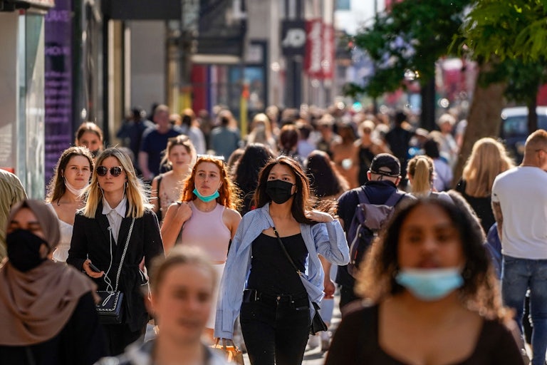 A crowd of people, some masked and some unmasked, walk down a busy street.