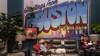 A masked woman sits on a couch at a furniture store turned warming station in front of a large sign reading “Greetings from Houston Texas”