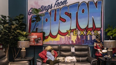 A masked woman sits on a couch at a furniture store turned warming station in front of a large sign reading “Greetings from Houston Texas”