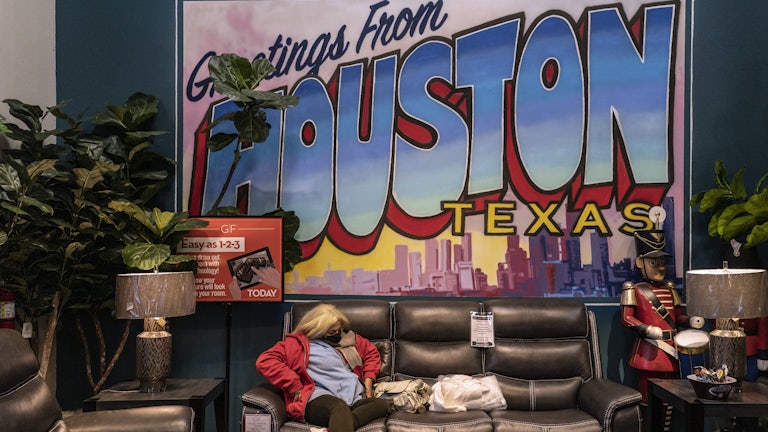 A masked woman sits on a couch at a furniture store turned warming station in front of a large sign reading “Greetings from Houston Texas”
