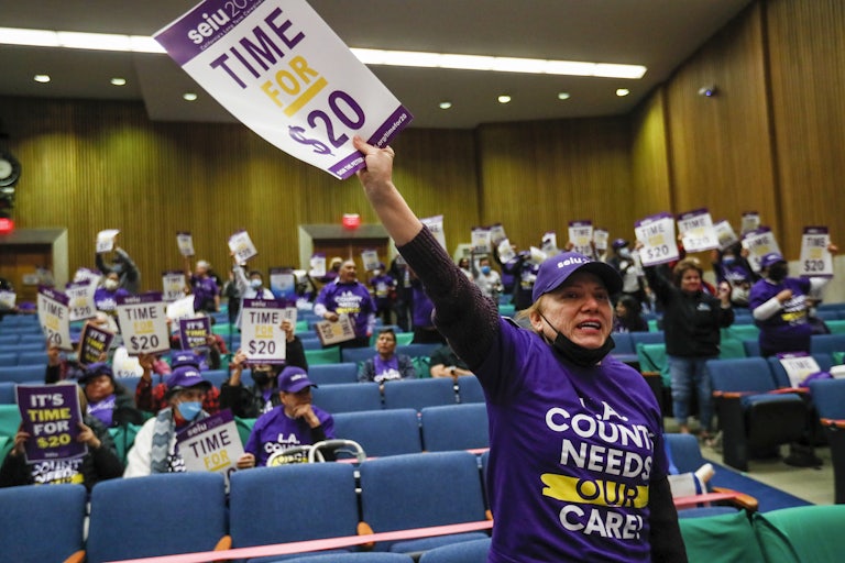 Workers wear purple tshirts that read "L.A. COUNTY NEEDS OUR CARE" and hold SEIU signs that read "Time for $20." A woman in the foreground raises her sign in the air and is chanting something.