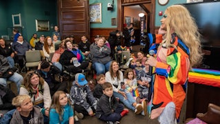 Children and adults sit and watch a drag performer standing with a microphone.