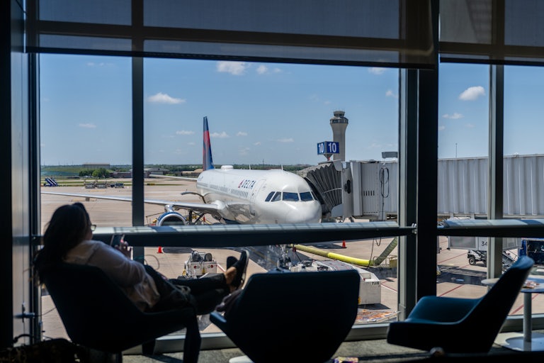 A person sits in a chair at an airport window, with a view of a plane on the runway.