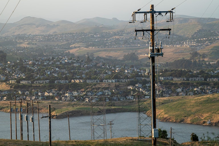 Power lines in the foreground are set against a neighborhood of homes on a hillside in the background. 