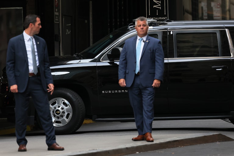 Two men in suits stand in front of a black SUV