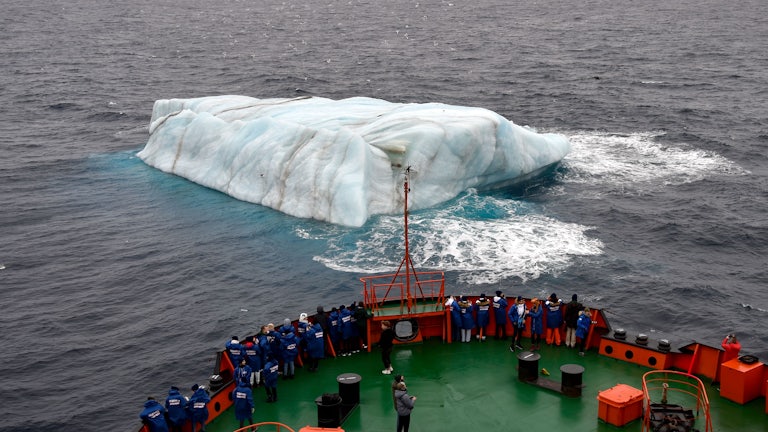 People stand on the deck of a ship, looking at an iceberg in the water.