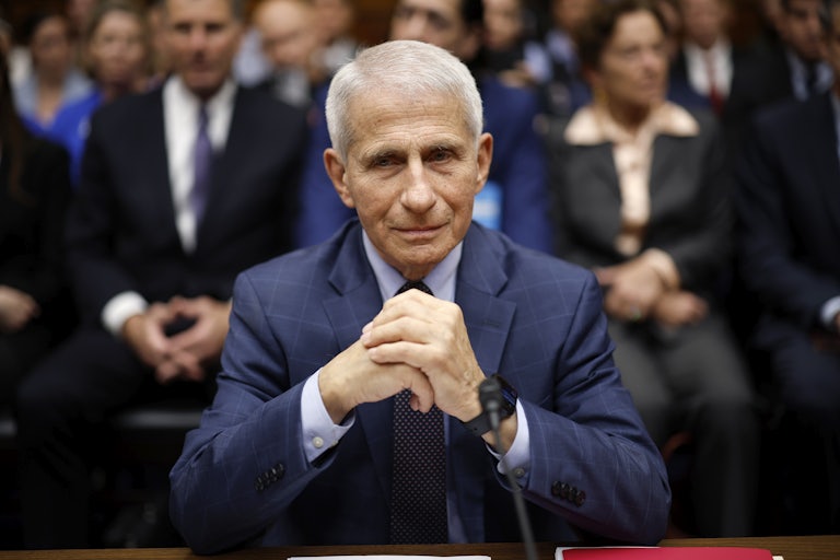 Anthony Fauci sits in front of a microphone with his hands folded