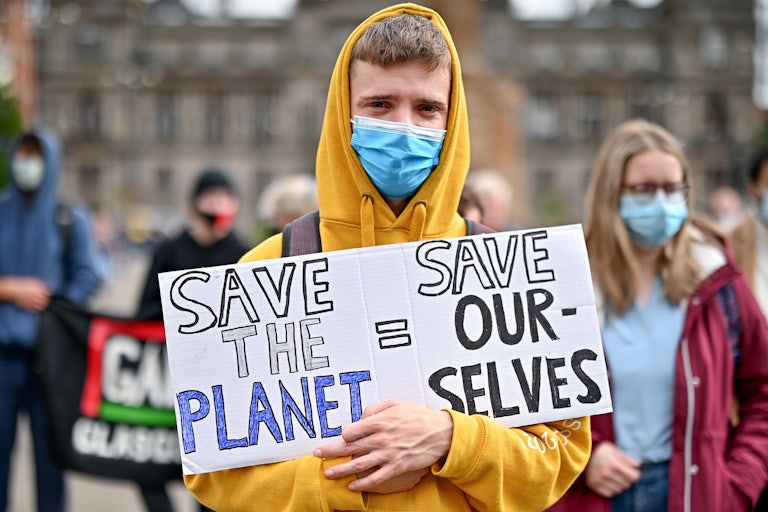 A young man in a hoodie holds a sign reading "save the planet = save ourselves."