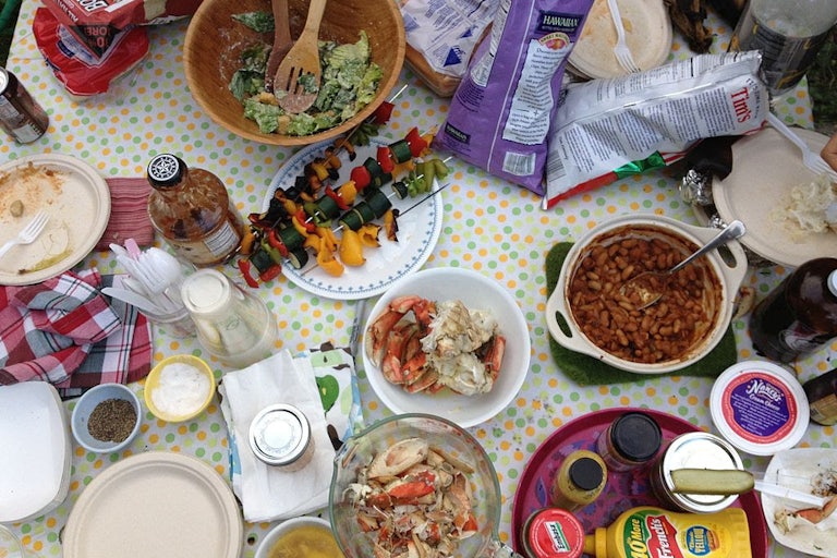 Dishes of crab, baked beans, vegetables on a skewer, salad, and condiments sit on a table alongside plastic cutlery and paper plates.
