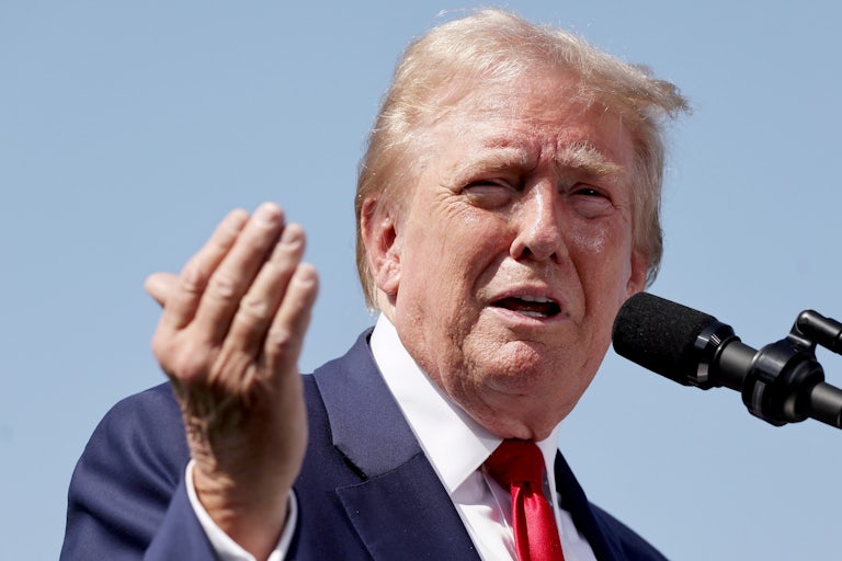 Donald Trump makes a weird face and a hand gesture while speaking into a mic