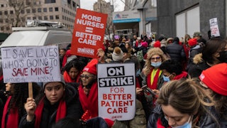 Striking health care workers hold up signs like "I SAVE LIVES WHO'S GOING TO SAVE MINE?," "ON STRIKE FOR BETTER CARE," and "MORE NURSES = BETTER CARE."