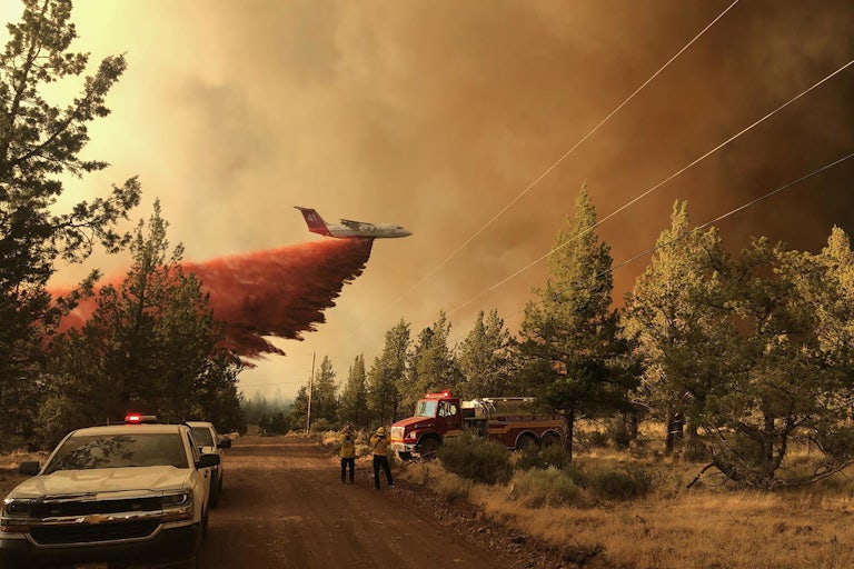An aircraft dumps fire retardant as emergency vehicles stand in the foreground.