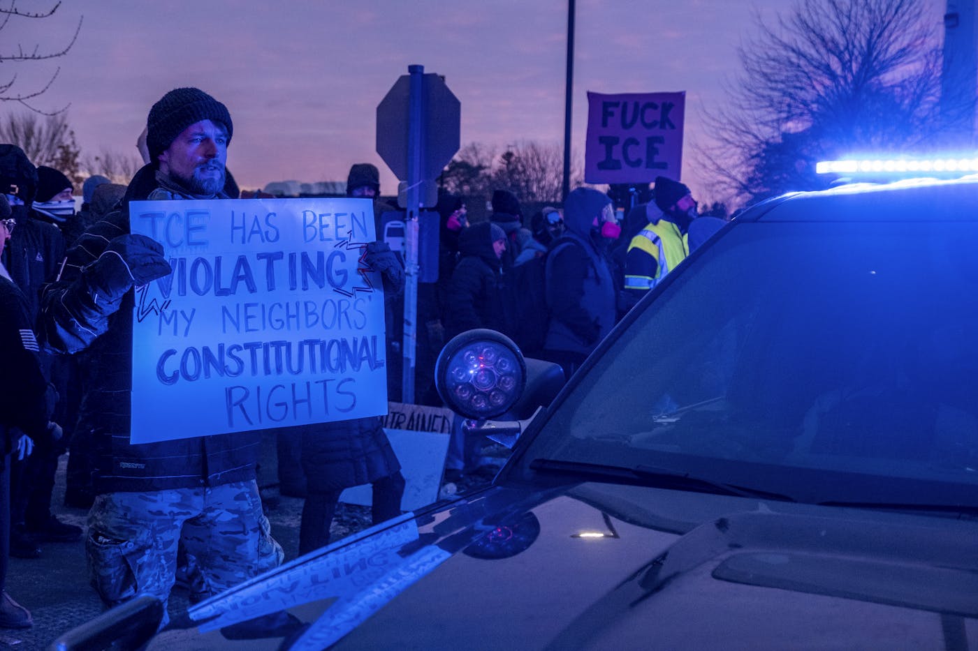 A protester holds a sign in front of a police vehicle that says ICE HAS BEEN VIOLATING MY NEIGHBORS CONSTITUTIONAL RIGHTS