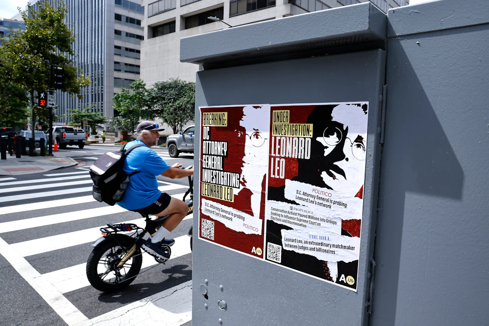 Two posters read "Breaking: D.C. attorney general investigating Leonard Leo" and "Under investigation: Leonard Leo."