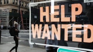 A woman is seen walking down the street in the reflection of a "Help Wanted" sign in a storefront window.