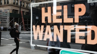 A woman is seen walking down the street in the reflection of a "Help Wanted" sign in a storefront window.