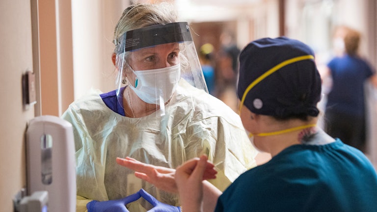 a nurse wearing a mask and face shield talks to another nurse in a Covid ICU