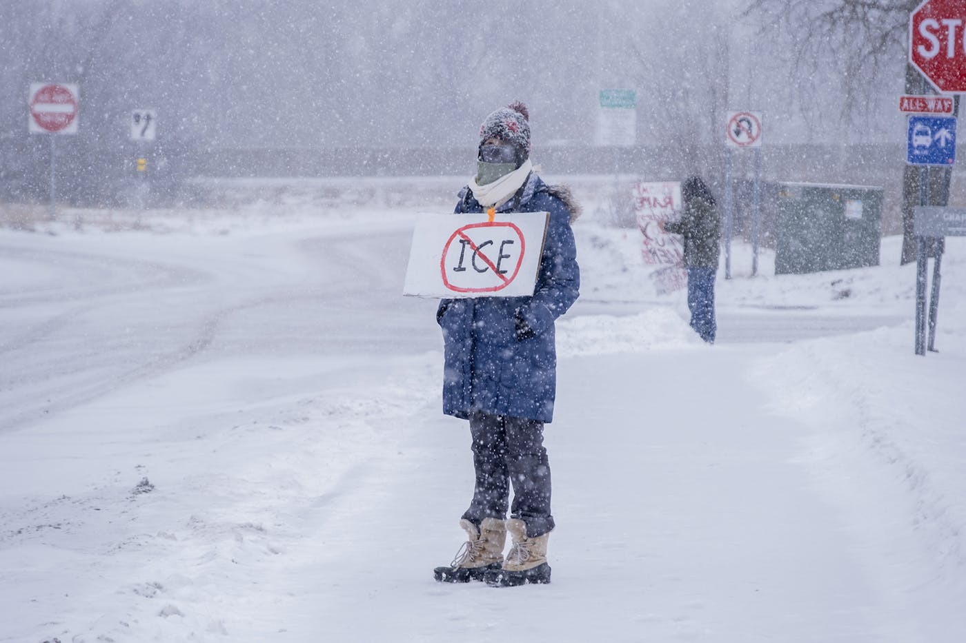 A woman stands in a blizzard by the intersection of a street with her hands in her pockets wearing a sign that says NO ICE