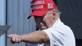 Donald Trump, wearing a red hat and a white polo shirt, points at a supporter and looks old.