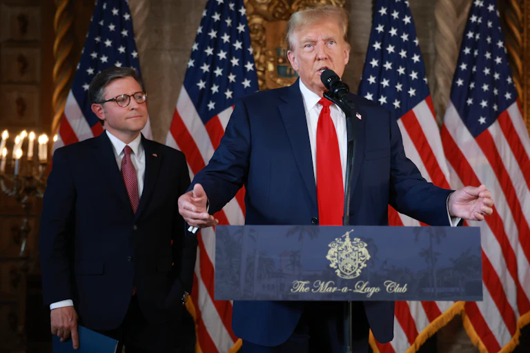 Donald Trump speak at a lectern while Mike Johnson stands behind him and looks on