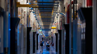 Two people walk down a corridor with columns of electronic equipment on either side. One pushes a cart.