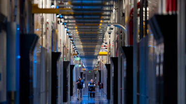 Two people walk down a corridor with columns of electronic equipment on either side. One pushes a cart.