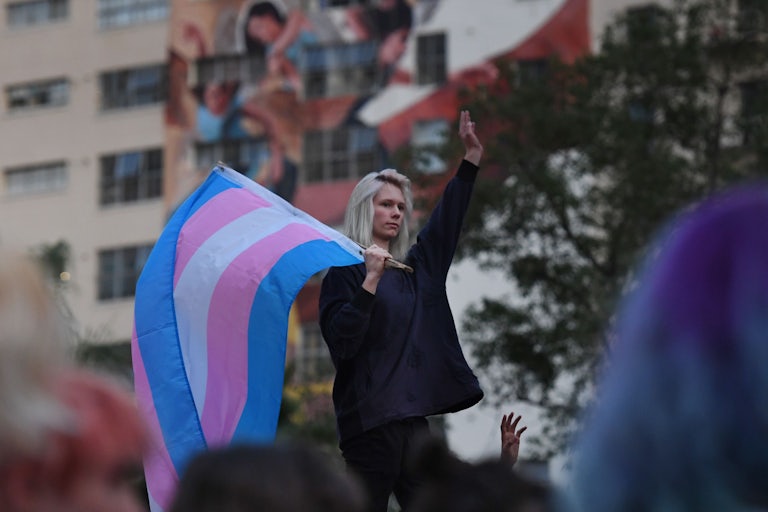 A protester holds a trans flag and raises their left hand in the sky.