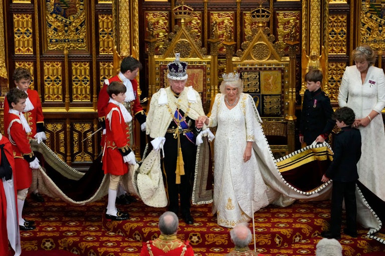 King Charles and Queen Camilla stand in ceremonial attire, surrounded by attendants holding their robes.