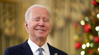 Joe Biden closes his eyes and does a wry little smile as he stands in front of a Christmas tree