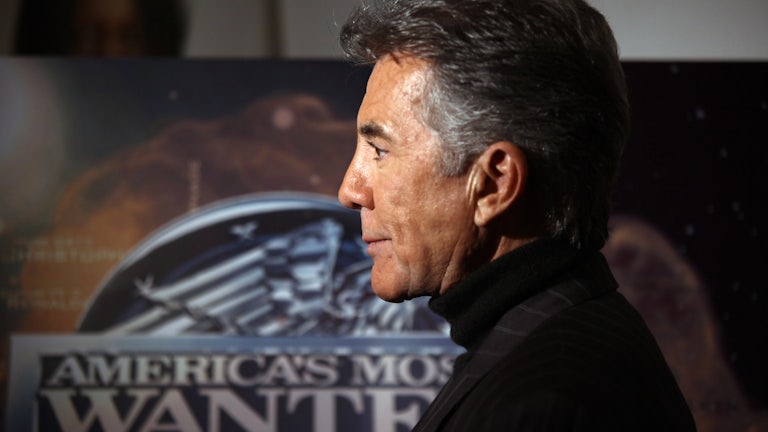 John Walsh, in profile, stands in front of a poster for “America’s Most Wanted”