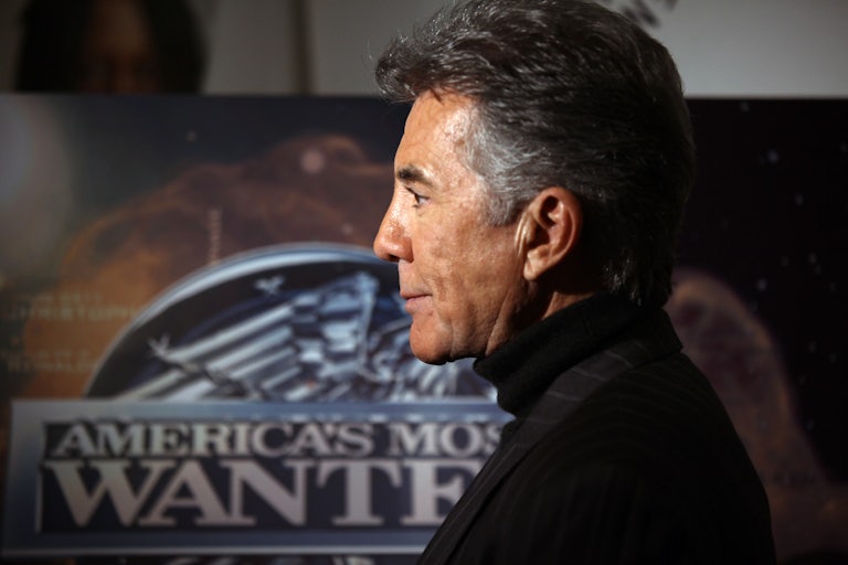 John Walsh, in profile, stands in front of a poster for “America’s Most Wanted”