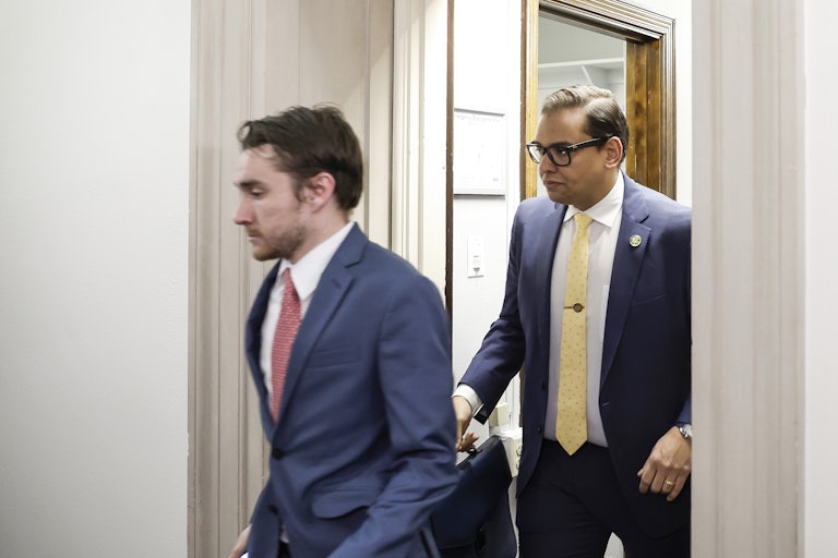 Representative George Santos walks through a doorway, briefcase in hand. A staff member walks ahead of him.