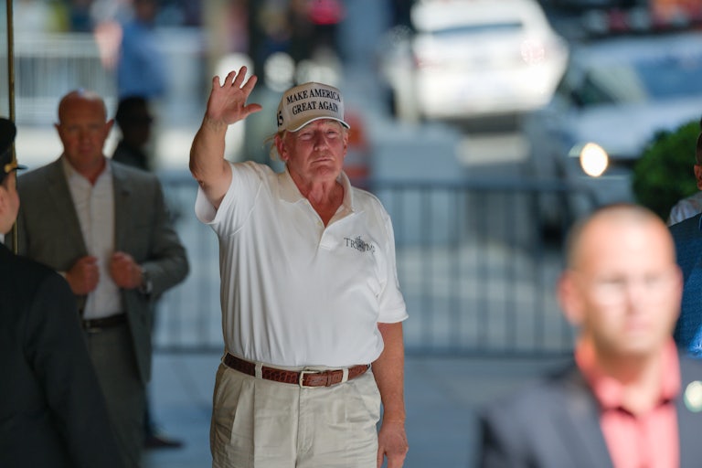 Donald Trump raises his hand in a wave. He wears a white Make America Great Again cap.