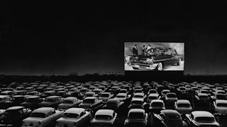 Vehicles fill a drive-in theater while people on the screen stand near a new car.