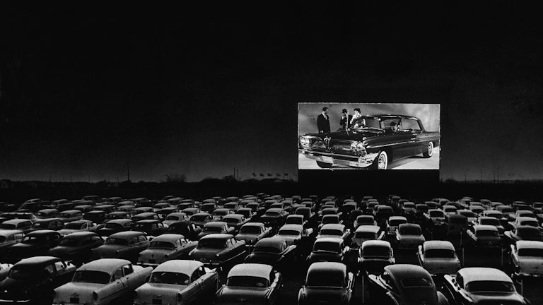Vehicles fill a drive-in theater while people on the screen stand near a new car.