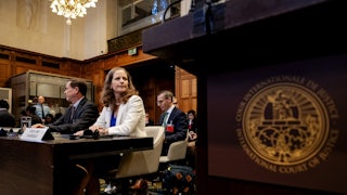 This photo, looking up from behind a lectern, shows a woman in a white blazer seated at a desk with a man in a suit to her right, with others in the room.